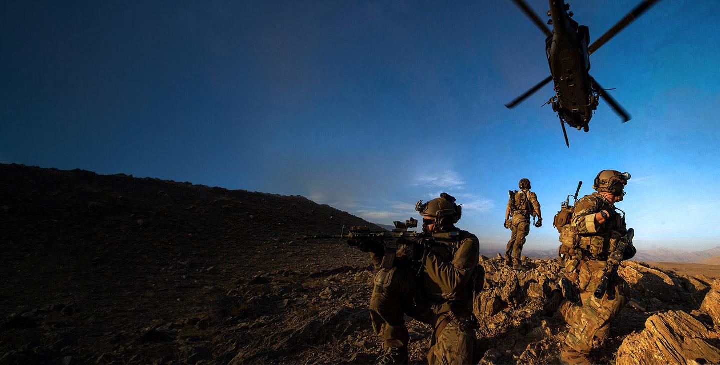 3名身着战斗装备的军人在一座多岩石的山上站岗，一架直升机在头顶飞过